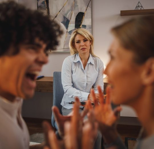 Shot of a couple having an argument during a counseling session with theirs female psychotherapist while being seated on a sofa inside of a living room.