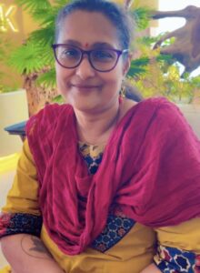 Smiling woman with purple glasses and red bindi wearing a yellow kurta and magenta scarf outdoors with greenery behind her